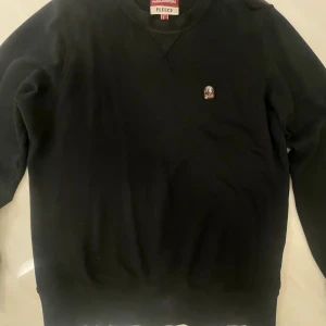 tröja Parajumpers L - Svart fleece-tröja från Parajumpers i storlek L. Tröjan har rund hals, lång ärm och ett broderat PJS-märke på bröstet. Ribbad nederkant med orange Parajumpers-tryck. Perfekt för dig som gillar stilren och bekväm streetwear.