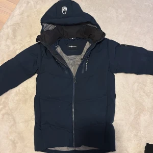 Mörkblå Sail Racing dunjacka med huva - Säljer en mörkblå dunjacka från Sail Racing med huva och snygga detaljer. Jackan har hel dragkedja, lång ärm och grått foder. Perfekt för kalla dagar och har en clean look med logga på ärmen och huvan. Pytte pytte pytte litet hål som inte syns det är på framsidan det syns knappt som ni ser på bilderna😃