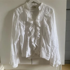 Vit blus med volanger - Supersöt vit blus med volanger längs knäppningen och spetsdetaljer framtill. Långärmad med lätt utsvängda ärmslut och v-ringad hals. Luftigt material som känns skönt mot huden och ger en romantisk vibe. Perfekt att styla med jeans eller kjol.