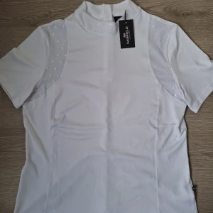 Vit ridtopp från Fairfield Equestrian - Snygg vit ridtopp från Fairfield Equestrian med korta ärmar och meshdetaljer med prickigt mönster på axlarna. Hög krage och stilren design, perfekt för dig som gillar hästar och vill ha en sportig look. Materialet är mjukt och följsamt. Stl M. Lappen kvar och aldrig använd och skickas i orginalförpackning!