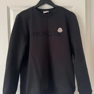Svart sweatshirt från Moncler - Svart sweatshirt från Moncler med broderad logga på bröstet och stort Moncler-tryck framtill. Tröjan har rund hals, långa ärmar och är gjord i mjuk bomull. Perfekt för dig som gillar stilrena och exklusiva plagg med streetkänsla.