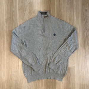 Grå half zip tröja från Timberland - Snygg grå tröja från Timberland med half zip och broderad logga på bröstet. Tröjan har långärmad design, ribbade muddar och är tillverkad i mjuk bomull. Perfekt för dig som gillar enkel och stilren look.