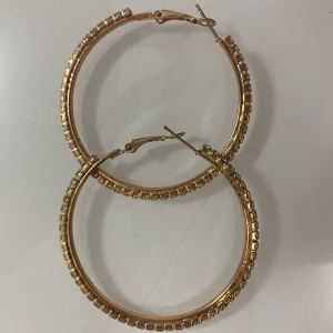 Guldiga örhängen  - Stora, runda örhängen i guldton med gnistrande stenar runt hela ringen. Klassiska med smidig låsning. Perfekta för att ge extra bling till din look.