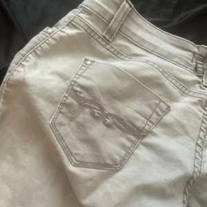 Beige jeans från Purlin Woman - Snygga beige jeans från Purlin Woman med klassisk femficksdesign och broderad detalj på bakfickorna. Byxorna har normal passform, bälteshällor och knappstängning framtill. Perfekta för dig som gillar stilrena och neutrala plagg.