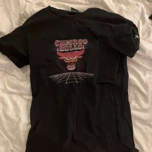 Chicago Bulls t-shirt från New Era - Svart t-shirt från New Era med Chicago Bulls-tryck på bröstet. Snygg grafisk design med röd och vit text samt bull-logga. Klassisk rund hals och korta ärmar, perfekt för dig som gillar basket och streetwear.