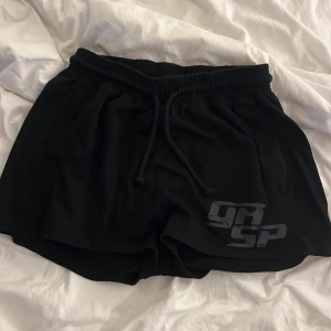 Svarta GASP shorts med tryck - Svarta shorts från GASP med elastisk midja och snörning. Tryck i grått framtill och guldigt GASP-logga baktill. Perfekta för gymmet eller chill. Mjukt och lätt material som känns skönt mot huden.