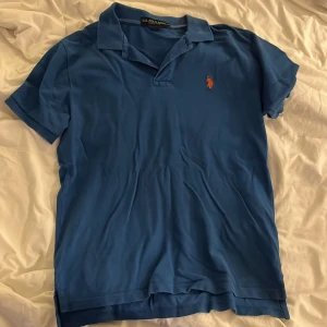 Blå pikétröja från U.S. Polo Assn. - Blå pikétröja från U.S. Polo Assn. med klassisk krage och korta ärmar. Tröjan har en orange broderad logga på bröstet och är tillverkad i 100% bomull. Perfekt för dig som gillar stilren och sportig look.