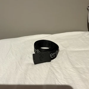 Svart Peak Performance bälte - Svart bälte från Peak Performance med vit logotyptext längs hela bandet. Bältet har en svart metallspänne med Peak Performance-logga. Tillverkat i slitstarkt syntetmaterial, perfekt för en sportig och avslappnad stil.