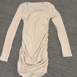 Beige ribbad kortklänning med rynk - Säljer en beige ribbad kortklänning med lång ärm och fyrkantig urringning. Klänningen har snygga rynkade detaljer längs sidan som ger en cool form och extra stilpoäng. Materialet är mjukt och stretchigt, perfekt för dig som gillar bekväma men trendiga plagg.