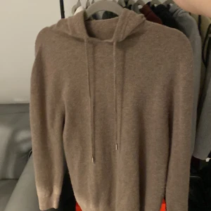 Beige hoodie i kashmir med snörning - Mjuk och lyxig beige hoodie i kashmir med huva och snörning framtill. Tröjan har långärmad design och en clean look utan synliga loggor eller mönster. Perfekt för dig som gillar stilrena och bekväma plagg med exklusiv känsla.