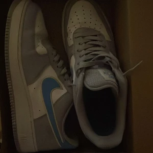 Nike Air Force 1 sneakers blå/grå - Säljer ett par Nike Air Force 1 i storlek 44. Klassisk modell i vitt och ljusgrått med ljusblå detaljer. Skorna är lite använda men i fint skick – inga större märken eller skador.