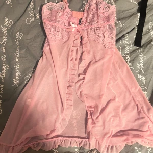 Rosa spetsklänning med volanger - Söt och feminin rosa klänning med spets upptill och volangdetaljer längs öppningen. Klänningen är kort och har en liten rosett framtill. Materialet är lätt och transparent, perfekt för dig som gillar romantisk stil.