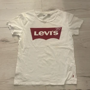 Vit Levi's t-shirt med röd logga - Vit t-shirt från Levi's i bomull med klassisk röd logga framtill. T-shirten har rund halsringning och korta ärmar. Enkel och stilren modell. Har dock en liten fläck se på sista bilden, går säkert bort i tvätt. 