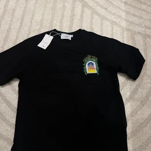 Svart t-shirt Casablanca S - Svart t-shirt från Casablanca Tennis Club i storlek S. Framsidan har ett litet färgglatt tryck med motiv av en byggnad och texten 'Avenida Casablanca', medan baksidan har ett stort, detaljerat tryck med samma motiv och gröna blad runt fönstret. Rund hals och klassisk passform.