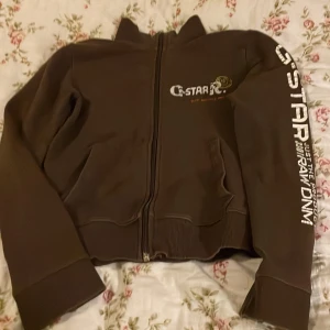 Brun långärmad från G-Star RAW - Snygg brun tröja med dragkedja  från G-Star RAW med vit logga på bröstet och längs ena ärmen. Tröjan  har ribbade muddar, två fickor fram och dragkedja. Materialet är mjukt och känns som bomull, perfekt för dig som gillar streetwear och vill sticka ut med coola detaljer.