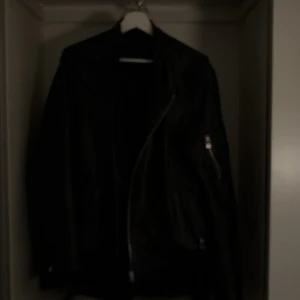 Svart skinnjacka med dragkedjor - Snygg svart skinnjacka med asymmetrisk dragkedja framtill och flera zip-detaljer på fickor och ärm. Jackan har klassisk bikerstil med krage och långärmade armar. Perfekt för dig som vill ha en cool och tidlös look.