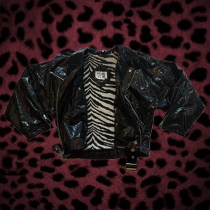 Svart glansig bomberjacka med zebra - Cool svart glansig bomberjacka med croppad passform och breda axlar. Insidan har ett fett zebramönster i svart och vitt. Jackan har flera dragkedjor, knappar och ett bälte i midjan. Perfekt för dig som vill sticka ut med edgy stil. Passar S/M