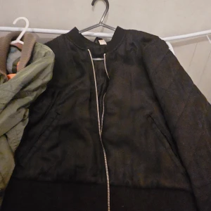 Svart bomberjacka med dragkedja - Snygg svart bomberjacka med klassisk rund krage och silverfärgad dragkedja framtill. Jackan har långa ärmar och ribbade muddar vid ärmslut och nederkant. Perfekt för dig som gillar en stilren och urban look.