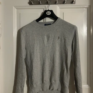 Grå tröja från Polo Ralph Lauren - Snygg grå tröja från Polo Ralph Lauren med klassisk rund halsringning och diskret broderad logga på bröstet. Tröjan är långärmad och har ribbade muddar vid ärmslut och nederkant. Perfekt för en clean och stilren look.