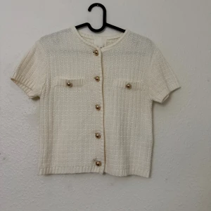 Vit stickad blus från H&M XS - Säljer en vit stickad blus från H&M i storlek XS. Toppen har korta ärmar, rund halsringning och dekorativa guldfärgade knappar framtill samt på de två bröstfickorna. Ribbad kant vid ärmslut och nederkant ger en snygg finish.