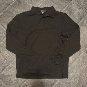 Svart långärmad piké från H&M - Svart ribbad piké från H&M med klassisk krage och knappar framtill. Långärmad modell i bomull med regular fit, perfekt för en clean och stilren look. Enkel att matcha med jeans eller chinos. Finns bild med på