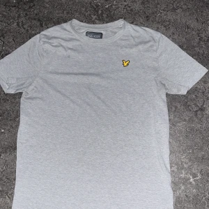 Ljusgrå t-shirt från Lyle & Scott - Snygg ljusgrå t-shirt från Lyle & Scott med klassisk rund hals och korta ärmar. På bröstet sitter den ikoniska gula fågeln som broderad detalj. T-shirten är gjord i mjuk bomull och har en clean, stilren look som funkar till det mesta.