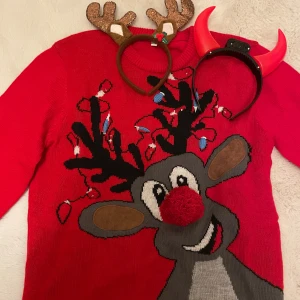 Röd jultröja med renmotiv - Rolig röd jultröja med stort motiv av en ren med julbelysning i hornen och en fluffig röd nos. Tröjan har rund hals och långa ärmar. Perfekt för att sticka ut på julfirandet!