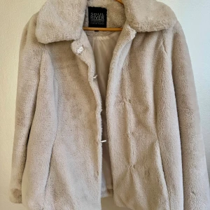 Beige pälsjacka från Soul River - Fluffig och mjuk beige (fusk)pälsjacka från Soul River med bred krage och diskret knäppning framtill. Jackan har en clean look och är helfodrad på insidan. Perfekt för dig som vill ha en cozy vibe och gillar neutrala färger.