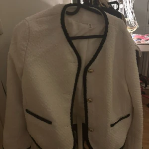 Vit cardigan jacka från H&M - Snygg vit cardigan jacka från H&M i storlek M men passar mig som vanligtvis har s ,Jackan har svarta kontrastkanter, guldfärgade knappar och två fickor framtill. Materialet är mjukt och strukturerat, perfekt för att lyfta din outfit med en klassisk touch.