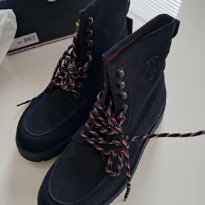 Tommy Hilfiger mörkblå mockaboots - Snygga mörkblå boots från Tommy Hilfiger i mjuk mocka med grov platt sula och rund tå. Skorna har coola flerfärgade snören och metallhål för snörning, samt röd insida och TH-logga på sidan. Perfekt för dig som gillar stilrena och robusta skor. Modellen heter: Tommy Hilfiger AMERICAN BOOT. 