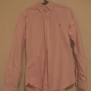 Rosa skjorta från Ralph Lauren M - Snygg rosa skjorta från Ralph Lauren i slim fit-modell. Klassisk design med knappar hela vägen, långärmad och med den ikoniska broderade loggan på bröstet. Skjortan är gjord i mjuk bomull och har en stilren krage som ger en clean look.