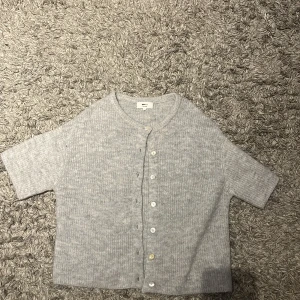 Grå stickad tröja från Gina Tricot - Säljer en ljusgrå tröja från Gina Tricot i mjuk ullblandning. Modellen är kortärmad med rund hals och knappar framtill. Perfekt för dig som gillar enkel och chill stil med lite struktur i materialet.