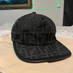 Svart Fendi keps med FF-mönster - Snygg svart keps från Fendi med ikoniskt FF-monogram över hela ytan. Kepsen har böjd skärm, justerbar rem bak med Fendi-logga och är tillverkad i bomull och polyester. Perfekt för dig som vill ha en lyxig och stilren accessoar.