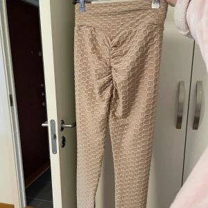Beige scrunch leggings med struktur - Säljer ett par beige leggings med scrunch-detalj bak och coolt rutmönstrat strukturtyg. Hög midja och tight passform, perfekta för att lyfta looken på gymmet eller till vardagsoutfiten. Storlek S-M