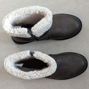 Bruna boots med fuskpäls från H&M - Säljer ett par bruna boots från H&M i storlek 36. De har en rund tå, platt sula och är fodrade med mjuk beige fuskpäls upptill. Bootsens ovandel är i syntetmaterial och de har en snygg spänndetalj på sidan samt dragkedja för enkel påtagning.