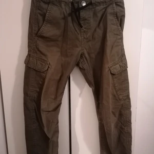 Olivgröna cargobyxor från KappAhl - Säljer ett par olivgröna cargobyxor från KappAhl med snörning i midjan och flera praktiska fickor på sidorna. Byxorna har en avslappnad passform och är tillverkade i bomull, perfekta för en chill och funktionell stil.