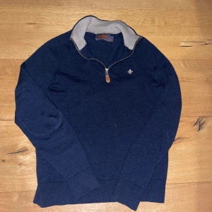 Marinblå stickad tröja från Morris - Snygg marinblå stickad tröja från Morris i 100% ull med halv dragkedja och kontrastfärgad krage i grått. Tröjan har långärmad design och en broderad logga på bröstet. Perfekt för dig som gillar klassisk stil med en modern touch. Tröjan har krympt så passar runt 145-155. Gör gärna också byten.