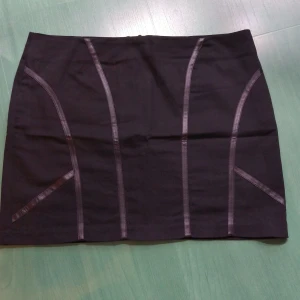 Svart kortkjol från b.p.c. bonprix - Svart kortkjol från b.p.c. bonprix med coola dekorativa sömmar framtill. Kjolen har en rak passform och är gjord i mjuk bomull med lite stretch för extra komfort. Stängs med dragkedja bak. Perfekt för dig som gillar stilrena och enkla plagg.