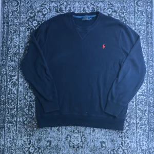 Mörkblå tröja från Polo Ralph Lauren - Snygg mörkblå tröja från Polo Ralph Lauren med klassisk röd broderad logga på bröstet. Tröjan har rund hals och långa ärmar, perfekt för dig som gillar stilrena och tidlösa plagg. Materialet är mjukt och känns skönt mot huden.