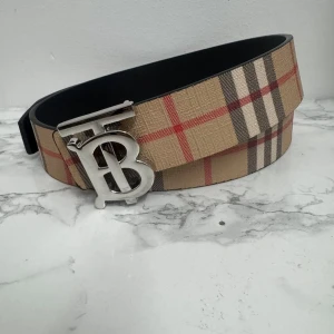 Burberry rutig bälte med silver-B spänne - Säljer ett snyggt Burberry bälte med klassiskt rutig design i beige, rött, svart och vitt. Bältet har ett brett band och ett stilrent silverfärgat spänne med Burberrys ikoniska B-logga. Perfekt för att lyfta din outfit med en lyxig touch.