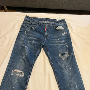 Levis blå slitna jeans med hål - Säljer ett par blå Dsquared2  jeans med slitningar, hål och färgstänk för en riktigt edgy look. Klassisk femficksmodell med rakt snitt och låg midja. Perfekta för dig som gillar en avslappnad och streetinspirerad stil.