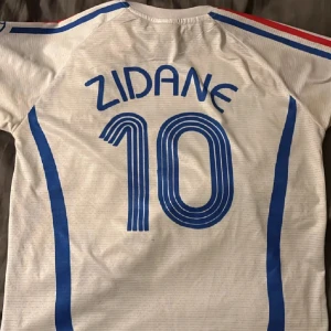 Zidane t shirt  - Säljer en vit Frankrike t shirt från Adidas med blå och röda detaljer. Tröjan har korta ärmar och nummer 10 på framsidan samt namnet 'Zidane' på baksidan. Från final 2006 Italien vs Frankrike. LIMITED EDITION! Haft den bara hängd uppe i rummet. Först till kvarn!