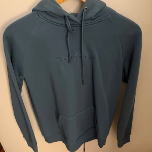 Blå hoodie - Peak Performance  - Snygg blå hoodie från Peak Performance i storlek L. Tröjan har broderad logga på bröstet, huva med snörning och en stor magficka. Perfekt för chill dagar och har en skön, mjuk känsla tack vare bomullsmaterialet. Är knappt använd och kommer ej till användning därav säljer💕