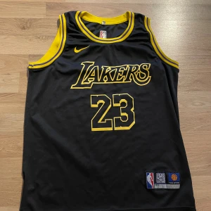 Svart Lakers basketlinne LeBron James - Snygg svart Lakers baskettröja med gula detaljer och nummer 23, LeBron James. Klassisk ärmlös modell i ventilerande meshmaterial, officiell NBA Team Apparel. Lakers-logga och Nike Swoosh framtill, namn och nummer bak.