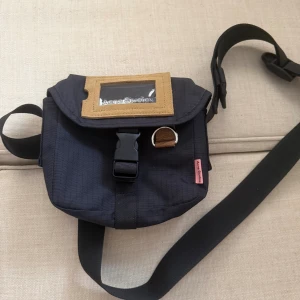 mini messenger väska från Acne Studios - Cool svart axelväska från Acne Studios i slitstark polyester med justerbar rem, spänne framtill och beige detalj med transparent fönster på locket. Praktisk och stilren design med diskret rutmönster i tyget och rosa logotagg på sidan.