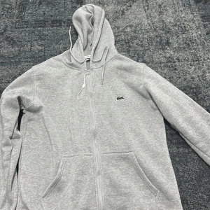 Grå hoodie från Lacoste med dragkedja - Snygg ljusgrå hoodie från Lacoste med klassisk krokodillogga på bröstet. Tröjan har dragkedja framtill, huva med snörning och två stora fickor. Tillverkad i mjuk bomullsblandning och har en relaxed passform som passar perfekt till chill dagar.