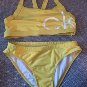 Gul bikini från Calvin Klein - Supersnygg gul bikini från Calvin Klein med sportig topp och klassiska bikiniunderdelar. Topp med vita CK-bokstäver framtill och coola dubbla axelband. Underdelen har Calvin Klein-logga baktill. I mycket bra skick men har små stygn som gått upp, men går att fixas! Som man kan se på sista bilden 💗