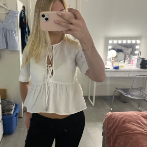 Vit blus med knyt från H&M - Supersöt vit blus från H&M med korta ärmar och två knyt framtill. Blusen har en rund halsringning, volang nedtill och är lätt croppad. Tillverkad i mjuk bomull som känns skön mot huden. Perfekt att styla med jeans eller kjol för en fräsch look.
