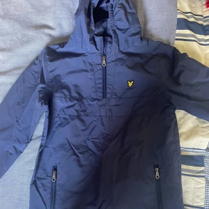 Marinblå windbreaker från Lyle & Scott - Snygg marinblå vindjacka från Lyle & Scott med huva och halv dragkedja framtill. Jackan har två sidofickor med dragkedja och den klassiska gula fågel-loggan på bröstet. Perfekt för blåsiga dagar och enkel att matcha med jeans eller joggers.