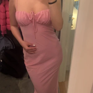 Rosa långklänning med smala axelband - Supersöt rosa långklänning med smala axelband och knytning framtill vid bysten. Klänningen har en tight passform och är ärmlös, perfekt för dig som gillar en feminin och enkel stil. Materialet känns mjukt och följsamt mot kroppen.